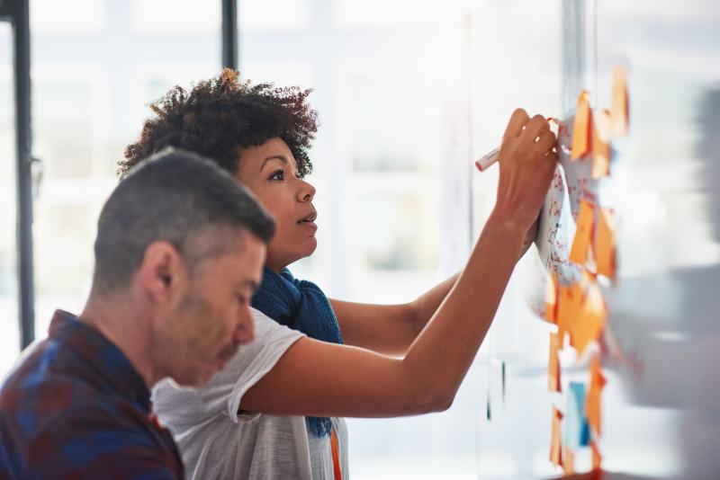Woman writing on a white board with sticky notes planning out strategy Woman writing on a white board with sticky notes planning out strategy
