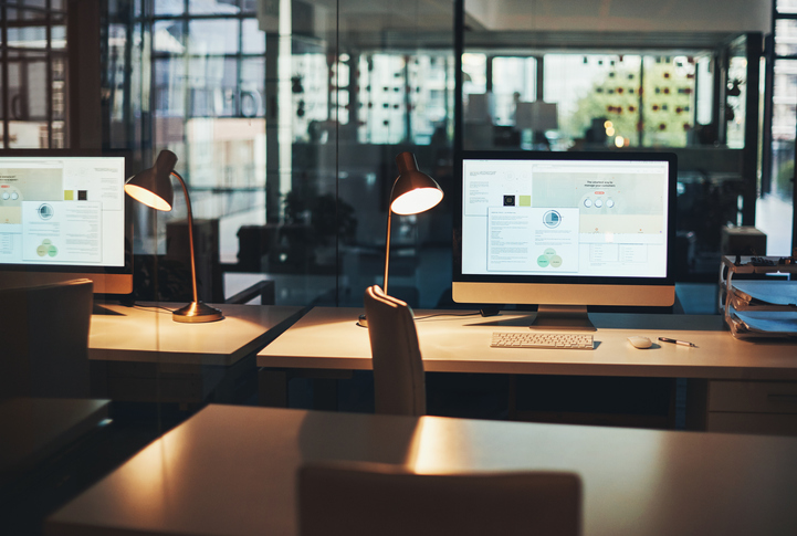 Two desktop monitors on a desk. The room is dim, but there is a single lamp. 