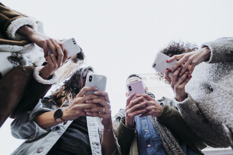 Four young adults standing in a circle looking at their phones,. Their faces are barely visible.