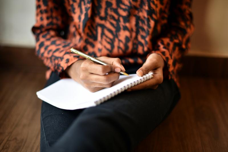 Woman from the shoulders down sitting on a bench with a pen and notebook in hand. She is wearing a patterned orange shirt.