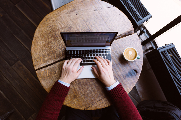 Bird's eye view of a man working on his laptop on a circular, wooden, coffee table with a cup of coffee.