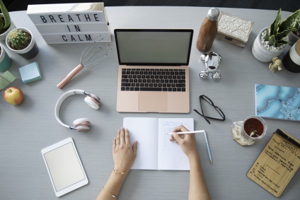 Person writing surrounded by laptop, headphones, notes, etc.