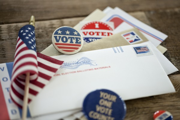 Pile of mail-in voting envelopes and "I Voted" stickers and an American flag