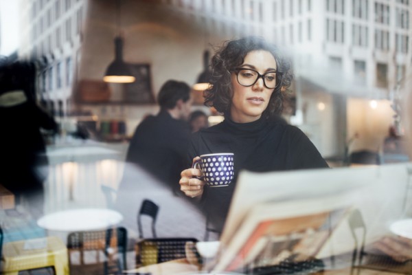 White woman with short hair and glasses sitting in a coffee shop looking at a newspaper with a cup of coffee in her hand.