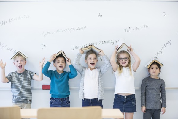 5 children standing against a white board with books on their heads. 